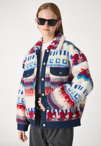 Chaqueta blanca difusa con patrones geométricos multicolores; puños y cuello de mezclilla azul; cierre frontal de botones; ajuste oversize.