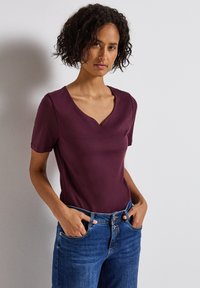 Burgundské tričko s krátkým rukávem a designem do V, vyrobené z měkké tkaniny, v kombinaci s modrými džíny. Střih je neformální a přiléhavý.