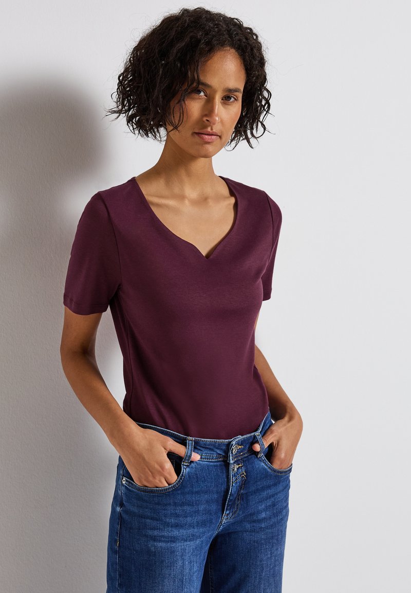 Burgundské tričko s krátkým rukávem a designem do V, vyrobené z měkké tkaniny, v kombinaci s modrými džíny. Střih je neformální a přiléhavý.