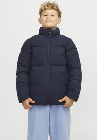 Marineblauer gepolsterter Jacke mit hohem Kragen, durchgehendem Reißverschluss und horizontalen Steppdetails. Getragen über hellblauen Jeans.