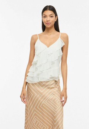 Kvinde iført en hvid lagdelt rynket camisole og et beige skørt med hvide prikker i geometriske mønstre, stående mod en ensfarvet baggrund.