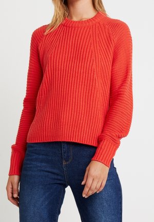 Femme portant un pull côtelé rouge vif à manches longues et un jean bleu foncé, debout devant un fond blanc uni.