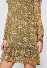 Robe en tissu transparent à fleurs vert olive avec manches longues bouffantes et ourlet à volants, portée par une personne dont la main est visible sur le côté.