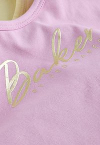 Tissu rose avec texte métallique doré "Baker by Ted Baker" imprimé dessus, montrant un gros plan sur l'encolure et la texture.