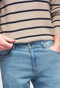 Pull beige à rayures bleu marine associé à un jean en denim bleu clair, avec des poches visibles et un bouton en métal à la taille.