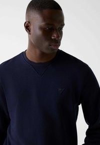 Sweat-shirt bleu marine avec un col rond, des poignets côtelés et un logo triangulaire sur la poitrine. Texture lisse, coupe décontractée.