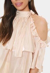 Pink, flowing blouse with ruffled cold shoulder design. Made from a shiny, lightweight fabric. Features a tie at the neck and soft pleats.