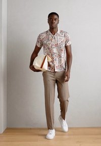 Kortærmet skjorte med geometrisk mønster i creme, rød og brun, parret med beige bukser og hvide sneakers, mens der bliver holdt en taske i creme og brun.