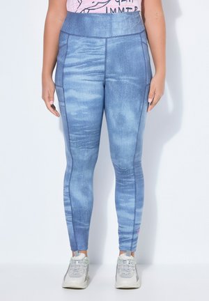 Frau trägt blaue Leggings mit hoher Taille und Denim-Struktur sowie weiße Sneakers, steht vor einem schlichten weißen Hintergrund.