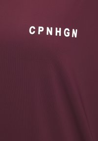 Burgundijas īsām piedurknēm krekls ar gludu tekstūru, uz kura priekšpuses centrā ir balti burti "CPNHGN". Vienkāršs dizains un regulāra piegriezums.