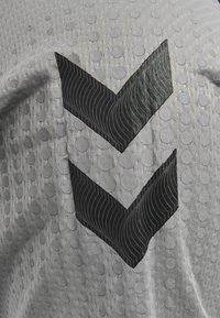 Grå tekstil med forhøjede cirkulære mønstre og to sorte chevron-designs med teksturerede accenter, der skaber kontrast til baggrunden.