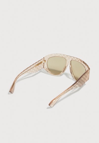 Lunettes de soleil beige transparent avec des verres légèrement teintés et un motif monogramme subtil sur les montures et les branches.
