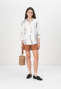 Witte bloemetjesprint shirt met driekwart mouwen, bruine shorts en een geweven handtas. Zwarte veterschoenen maken de outfit compleet.