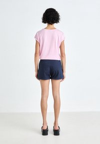 Kvinna med lockigt svart hår, iklädd en rosa och vit randig kortärmad topp, marinblå shorts med uppvikta ben och svarta platåsandaler, stående mot en vit vägg.