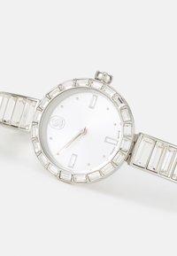 Silver watch featuring a round face, minimalist hour markers, and a crystal-accented bezel. The band has rectangular links and a shiny finish.