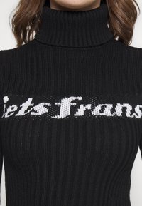 Czarny dziergany sweter z golfem o prążkowanej fakturze i białym kursywnym napisem „Lets France” na wysokości klatki piersiowej.