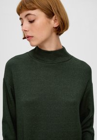 Ciemnozielony sweter z wysokim kołnierzem, o miękkiej fakturze, luźnym kroju, długich rękawach, minimalistycznych szwach, gładkim wzorze dzianiny oraz bez widocznych elementów metalowych czy akcentów.