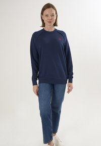 Felpa blu navy con maniche raglan e logo a cuore rosso sul lato sinistro del petto, abbinata a jeans a gamba dritta azzurri e sneakers bianche.