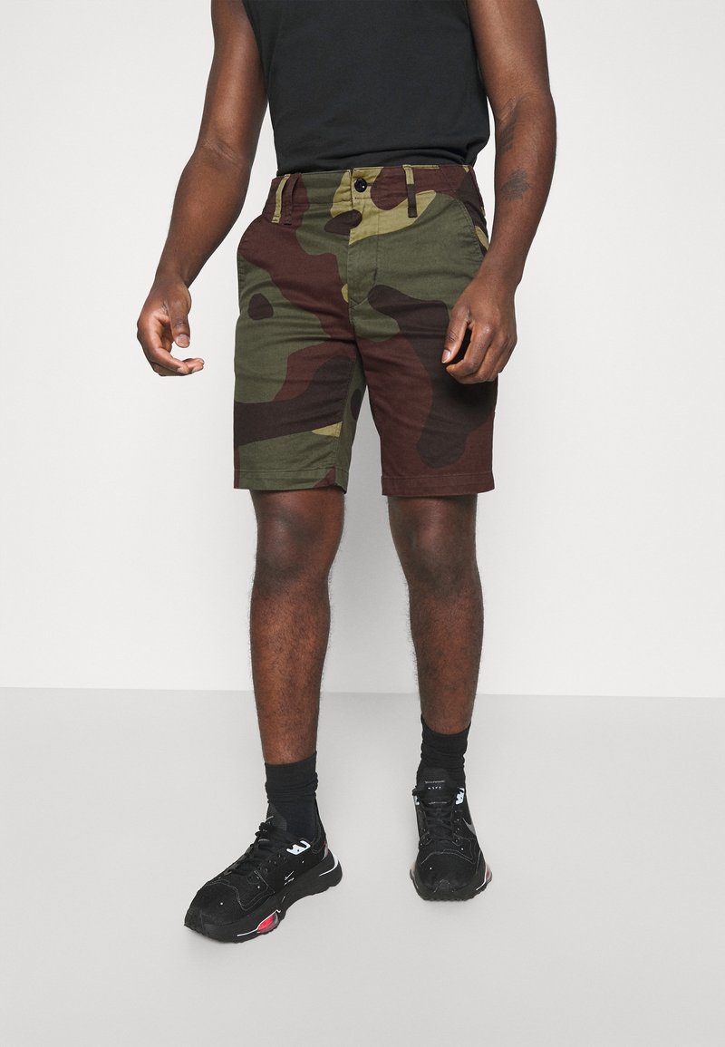 Shorts camouflage avec des motifs verts, marron et noirs. En coton ; dotés de poches et d'une coupe droite. Portés avec des chaussures de sport noires.
