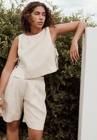 Haut sans manches en lin beige avec des liens sur les côtés, associé à un short plissé assorti, présentant une coupe décontractée et une finition texturée.