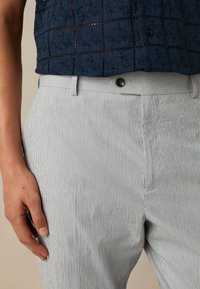 Pantalon à rayures verticales de couleur claire avec une texture lisse, doté d'une fermeture à boutons et de poches latérales. Un design ajusté.