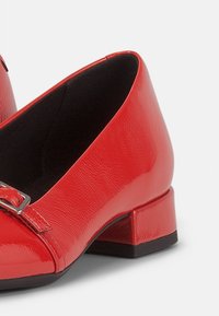 Mocasines de charol rojo con un tacón cuadrado bajo, que presentan un detalle de hebilla metálica en la parte frontal y un acabado suave y brillante.
