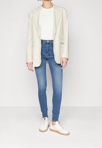 Ljusbeige oversized blazer, tajt vit topp, blå skinny jeans och vita sneakers med beige sulor. Släta texturer, avslappnad stil.