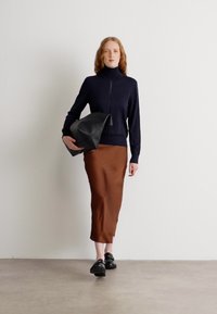 Marineblå høyhalset genser, brun satengskjørt og svarte sko. Modellen holder en stor svart toteveske. Glatte teksturer og elegante silhuetter.