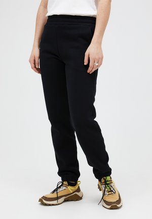 Person iført sorte tilspidsede bukser, lysebrune og brune sneakers samt en hvid top, vist fra taljen til fødderne mod en ensartet hvid baggrund.