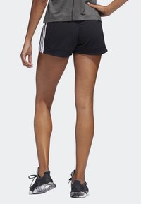 Pantalones cortos deportivos negros con rayas blancas en los laterales, combinados con zapatillas negras con suela texturizada. La vista trasera muestra un ajuste holgado y longitud.