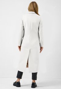 Manteau blanc sur mesure avec un col cranté, une fente au dos et des poignets boutonnés, associé à un pantalon noir cropped et des chaussures noires chunky.