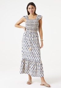 Maxikleid in Creme mit blauer Diamantmuster, mit Rüschäffchen, gesticktem Oberteil und rüschenförmigem Saum. Verstellbarer Kordelzug in der Taille mit Quasten.