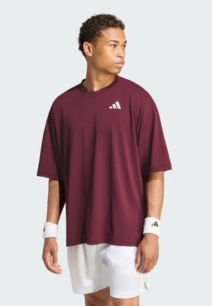 Junger Mann mit locker sitzendem dunkelrotem T-Shirt, weißen Shorts und weißen Handgelenkbandagen, der steht und seitlich schaut vor hellem Hintergrund.