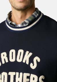 Námořnický svetr s krémovým nápisem "BROOKS BROTHERS." Pletená textura, žebrovaný límec s námořnickými a krémovými pruhy, překrytý límcem kostkované košile.