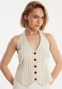 Beige Haltertop mit vertikalen schwarzen Streifen, tiefem V-Ausschnitt und einer Knopfleiste mit fünf schwarzen Knöpfen; glatte Stofftextur.