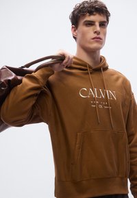 Brun hoodie med framficka och en vit logotyp på bröstet. Dragband, mjuk textur och ribbade muddar kompletterar designen.