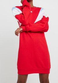 Rotes Sweatshirtkleid mit langen Ärmeln, lockerer Passform, gerippten Bündchen und blau-weißen Farbblockakzenten an den Schultern.