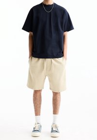 Homme portant un T-shirt bleu marine, un short beige à cordon, des chaussettes blanches et des baskets bleues, se tenant les mains dans les poches sur fond blanc.