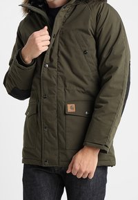 Olivově zelená zateplená bunda s černou podšívkou, s kapucí lemovanou kožešinou, několika kapsami a čtvercovou nášivkou loga na přední straně.