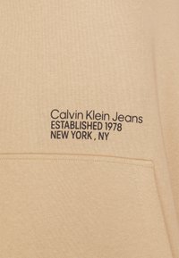 Hoodie beige avec une poche frontale et un texte brodé noir : "Calvin Klein Jeans, ÉTABLI EN 1978, NEW YORK, NY." Texture de tissu doux.