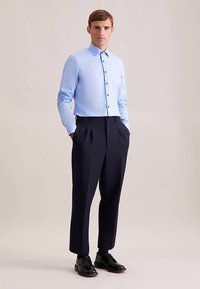 Lichtblauw overhemd met lange mouwen, gecombineerd met donkerblauwe geplooide broek. Model draagt zwarte leren nette schoenen.