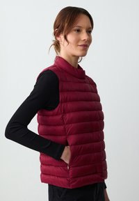Gilet puffa bordeaux con colletto alto, motivo trapuntato orizzontale e tasche con zip, indossato sopra un top a maniche lunghe nero.