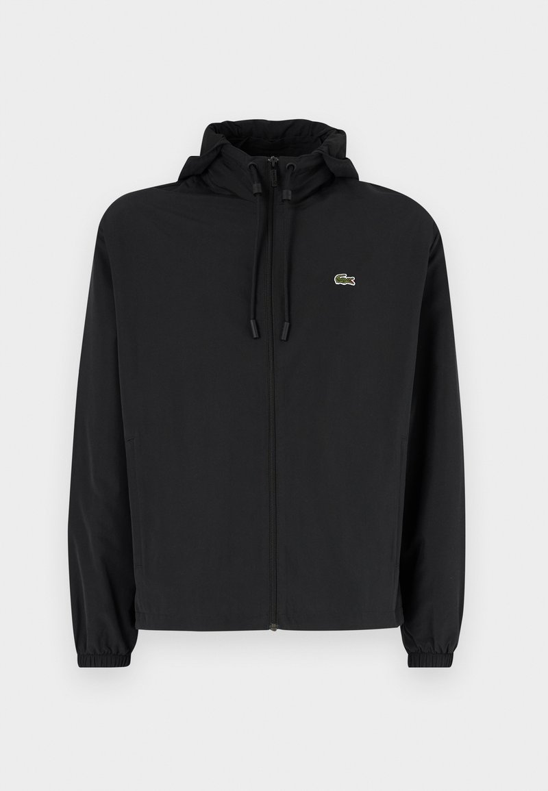 Veste noire à fermeture éclair avec capuche, fabriquée en matériau léger. Présente des poignets élastiques et un petit logo vert sur la poitrine gauche.