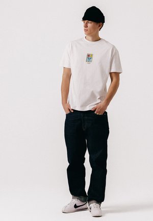 Hombre joven con gorro negro, camiseta blanca con un diseño de tulipán, vaqueros oscuros y zapatillas blancas Nike, de pie con las manos en los bolsillos.