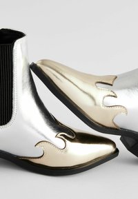 Botines metálicos en plata y oro con un diseño de punta afilada, paneles laterales de pana negra texturizada y acentos en forma de llama.