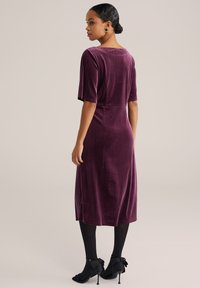 O femeie stând de profil, purtând o rochie din catifea violetă de lungime medie, colanți negri, pantofi cu toc înalt negri cu fundițe, cercei aurii și o colier.