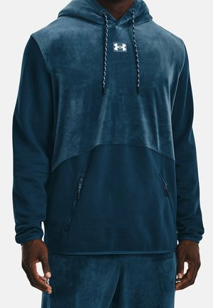 Mann trägt dunkelblaue Under Armour Kapuzenjacke und passende Hose mit Reißverschlusstaschen, steht vor einfachem Hintergrund.