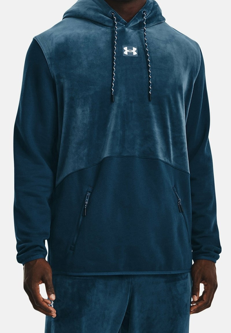 Homme portant un sweat à capuche Under Armour bleu foncé et un pantalon assorti avec des poches zippées, debout devant un fond uni.