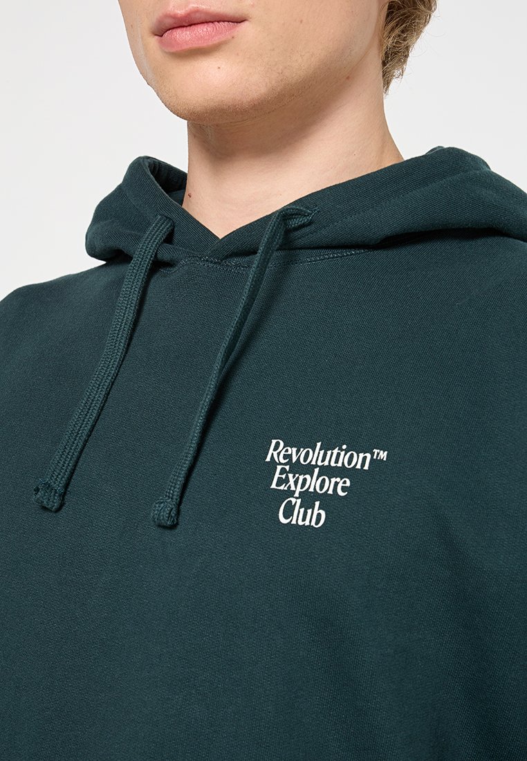 Mörkgrön hoodie med dragsko, med en vit textlogo "Revolution Explore Club" på framsidan. Tillverkad av mjukt, texturerat tyg.