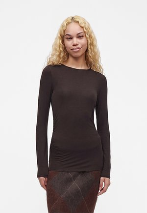 Młoda kobieta z długimi kręconymi blond włosami, ubrana w dopasowany, ciemnobrązowy top z długim rękawem oraz teksturowaną, ciemnobrązową spódnicę na jednolitym tle.